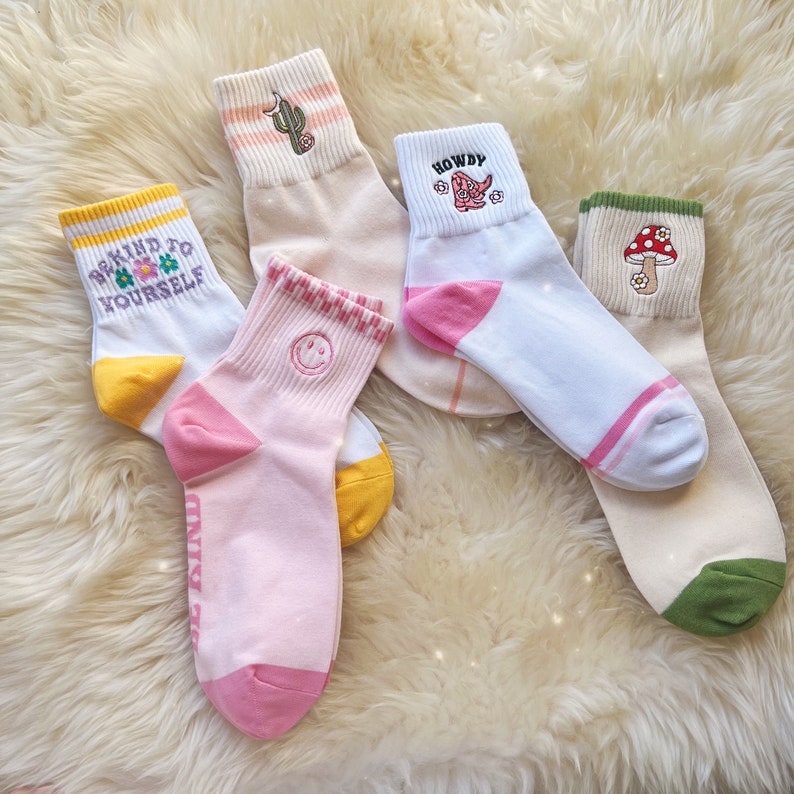 K&ouml;nnte beinhalten: F&uuml;nf Paar bunte Socken mit verschiedenen Designs. Die Socken zeigen einen Kaktus, einen Cowboystiefel, einen Pilz, ein Smiley und den Text "Be Kind To Yourself".