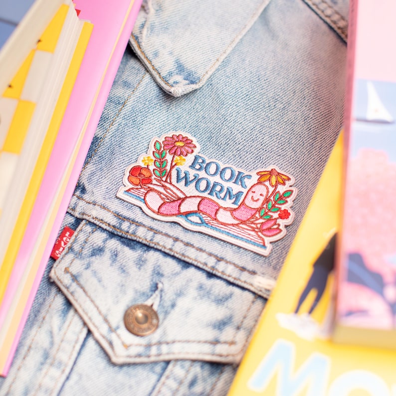 May include: A denim jacket with a colourful embroidered patch that reads "BOOK WORM." The patch features a smiling pink worm with flowers and leaves. Books in pink and yellow are in the background.