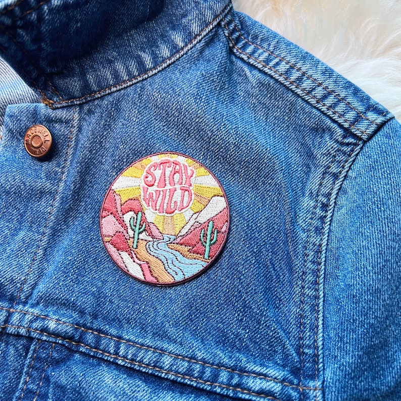 Peut inclure: Un &eacute;cusson brod&eacute; rond avec l'inscription "Stay Wild" dans un style r&eacute;tro, repr&eacute;sentant un paysage d&eacute;sertique avec des cactus, des montagnes et une rivi&egrave;re. L'&eacute;cusson est fix&eacute; sur une veste en jean bleue.