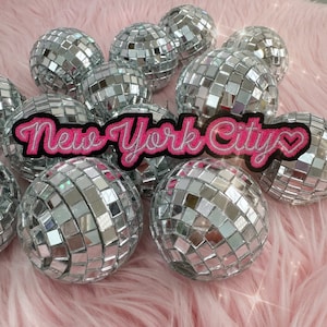 May include: A pink and white iron-on patch with the text "New York City" and a heart. The patch is surrounded by silver disco balls.