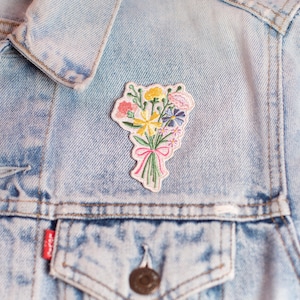 May include: Embroidered patch featuring a colorful bouquet of flowers tied with a pink ribbon. The patch includes yellow, pink, blue, and purple flowers with green stems and leaves. The patch is on a light blue denim jacket.