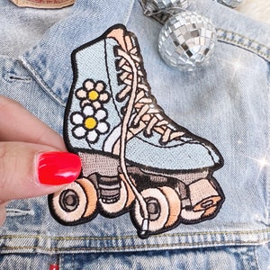 May include: Embroidered patch of a light blue roller skate with daisy details. The patch has tan laces and wheels, set against a denim jacket. A small disco ball is in the background.