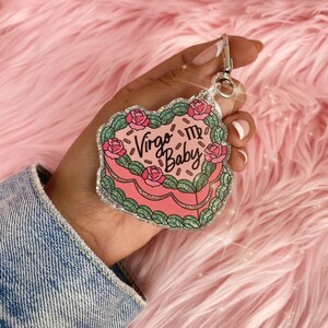 May include: Pink glitter heart-shaped keychain with the text "Virgo My Baby" and a Virgo zodiac symbol. The keychain is decorated with green leaves and pink roses.