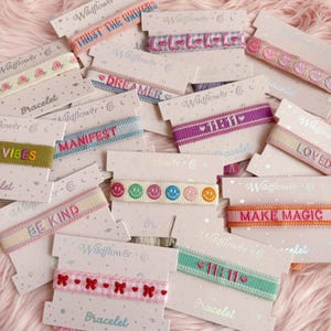 May include: Assortment of colorful woven bracelets with positive affirmations and symbols. The bracelets feature phrases like "Trust the Universe", "Dreamer", "Manifest", "Be Kind", "Make Magic", "Loved", "Vibes", and "11:11". The bracelets are packaged on cards with the Wildflower logo.