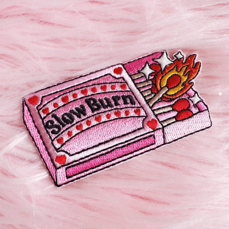 Puede incluir: Parche bordado rosa con dise&ntilde;o de caja de cerillas y texto "Slow Burn". La caja est&aacute; decorada con corazones y estrellas rojas, y se ve una cerilla encendida con llamas. Para ropa o accesorios.