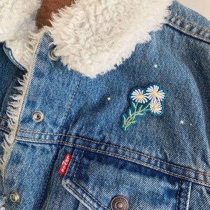 May include: A blue denim jacket with a white faux fur collar and a white daisy flower patch.