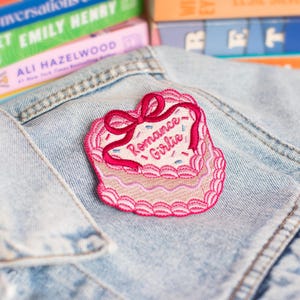 Può includere: Una toppa ricamata rosa a forma di torta con un fiocco rosso e le parole "Romance Girlie". La toppa è su una giacca di jeans azzurra. I libri sono visibili sullo sfondo.