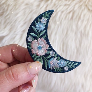 May include: A crescent moon shaped embroidered patch with a floral design. The moon is dark blue with green leaves and pink and blue flowers.
