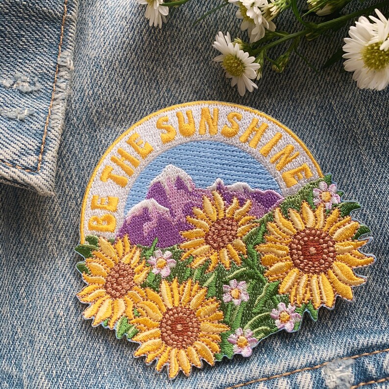May include: A yellow embroidered patch with the text "Be the Sunshine" in a white circle. The patch features a purple mountain range and three yellow sunflowers.