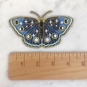 以下が含まれることがあります： 月と星のデザインが施された、青と金の刺繍の蝶のワッペン。ワッペンは約5cm幅です。