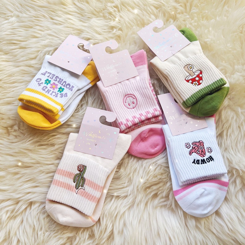 K&ouml;nnte beinhalten: Sechs Paar wei&szlig;e Baumwollsocken mit bunten Stickereien. Die Designs beinhalten Blumen, ein Smiley, einen Pilz, einen Kaktus und ein Schwein. Die Socken haben rosa und gr&uuml;ne Streifen.