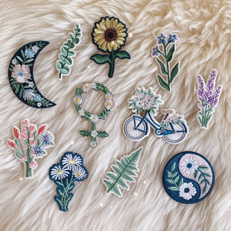 May include: A collection of embroidered iron-on patches featuring various designs, including a bicycle, a sunflower, a crescent moon, a fern, a yin-yang symbol, and several floral arrangements.