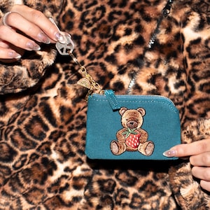 May include: Teal coin purse with a brown teddy bear embroidered with a strawberry. The purse has a gold zipper and a gold keychain with a rainbow charm. The background is a leopard print jacket.