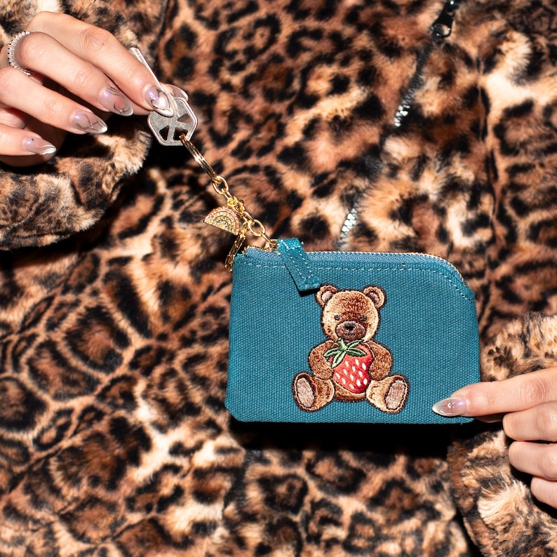 May include: Teal coin purse with a brown teddy bear embroidered with a strawberry. The purse has a gold zipper and a gold keychain with a rainbow charm. The background is a leopard print jacket.