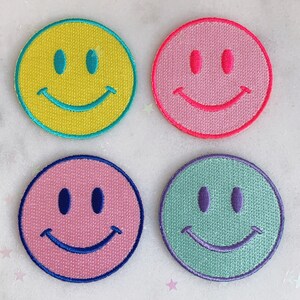 Puede incluir: Cuatro parches bordados con caritas sonrientes en diferentes colores: amarillo, rosa, azul claro y verde. Cada parche tiene un dise&ntilde;o simple de cara sonriente cosida.