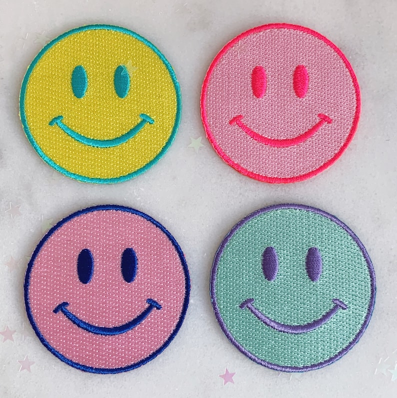 Puede incluir: Cuatro parches bordados con caritas sonrientes en diferentes colores: amarillo, rosa, azul claro y verde. Cada parche tiene un dise&ntilde;o simple de cara sonriente cosida.