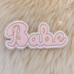 May include: Pink felt patch with gold stitching that reads "Babe" in a retro font.