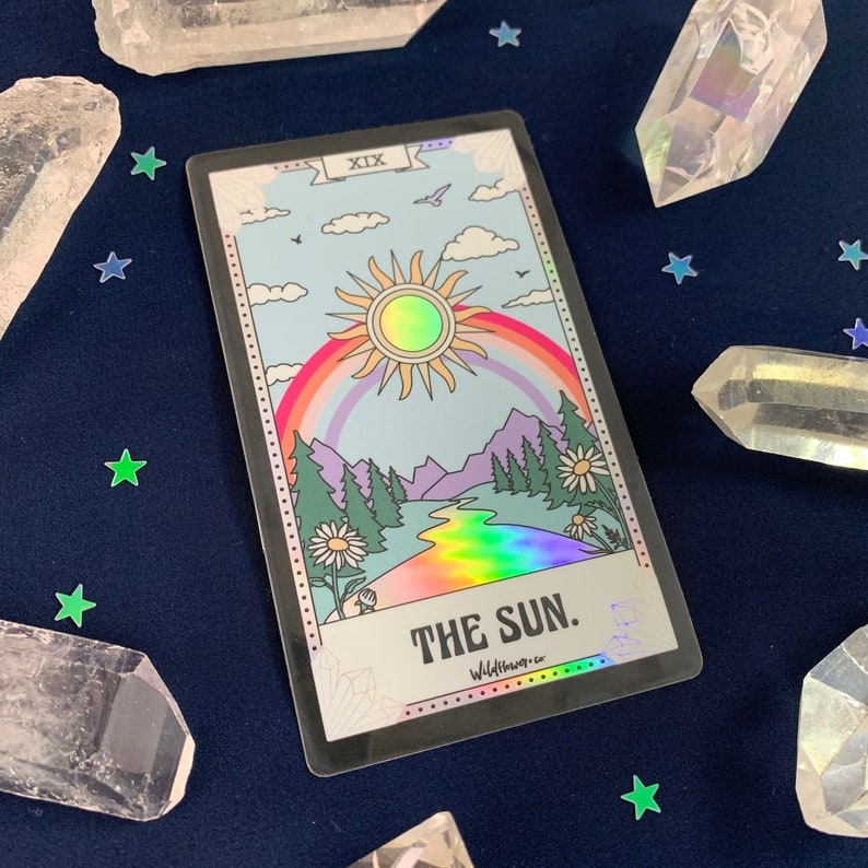 Puede incluir: Una carta de tarot hologr&aacute;fica con un arco&iacute;ris y un sol en un cielo azul. La carta est&aacute; etiquetada como "XIX. EL SOL." y "Wildflower Co."