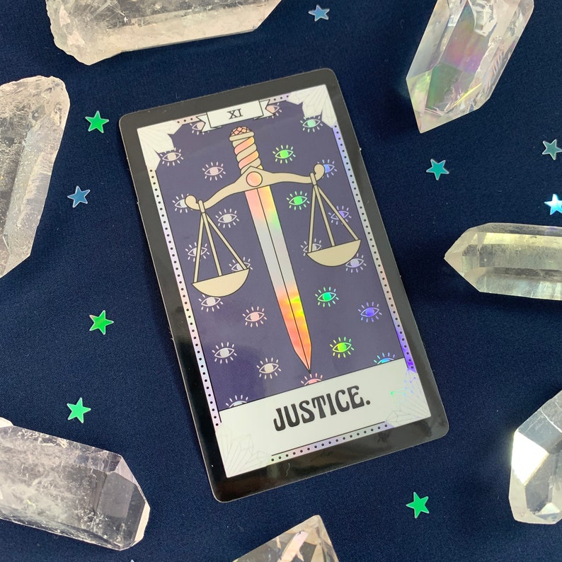 May include: A tarot card with a black background and a holographic finish. The card depicts a sword and scales, with the word "JUSTICE" printed below. The card is surrounded by a pattern of eyes.