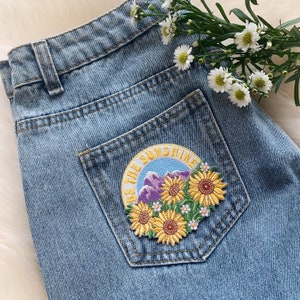 May include: A blue denim jean jacket with a patch on the pocket that says "Be the Sunshine" with a mountain and flower design.