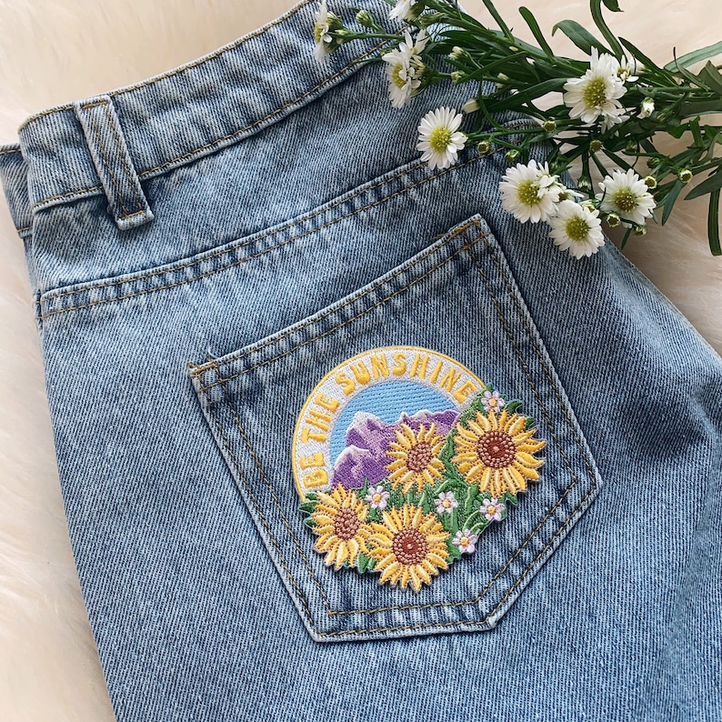 May include: A blue denim jean jacket with a patch on the pocket that says "Be the Sunshine" with a mountain and flower design.
