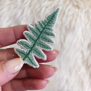 May include: A green embroidered fern leaf patch with white stitching around the edges.