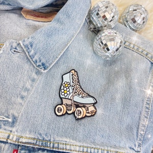 May include: A light blue roller skate patch with daisy accents is attached to a denim jacket. The patch has a retro design with cream-coloured wheels and laces. Three disco ball ornaments are in the background.