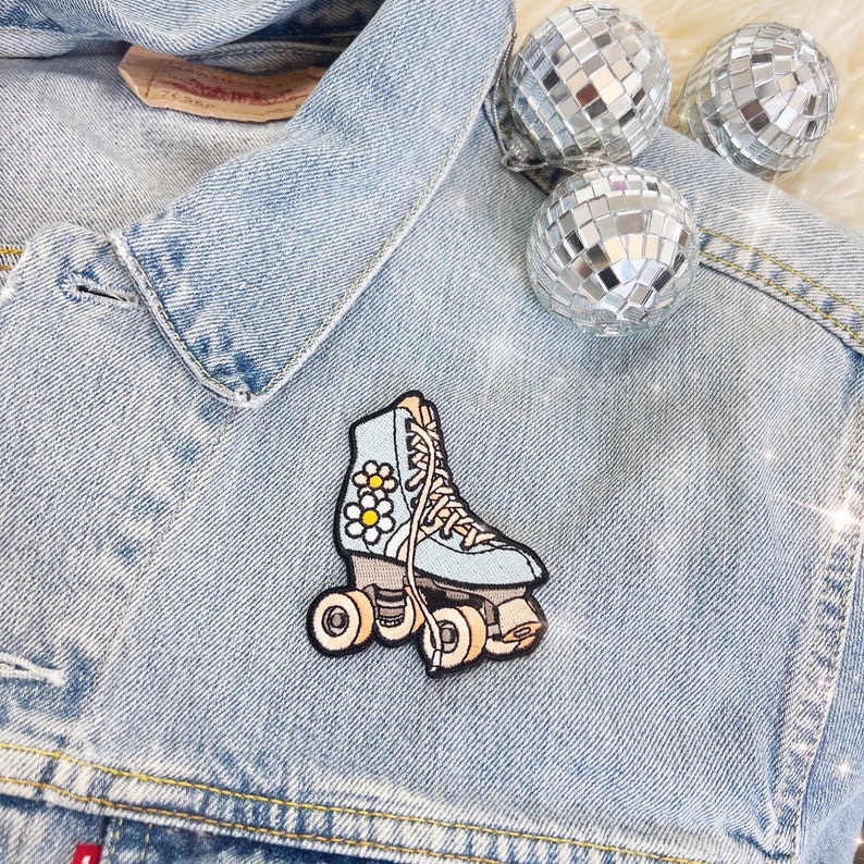 May include: A light blue roller skate patch with daisy accents is attached to a denim jacket. The patch has a retro design with cream-coloured wheels and laces. Three disco ball ornaments are in the background.