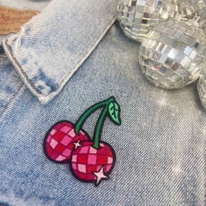May include: Embroidered cherry patch with a green stem and leaf, and pink and red checkered cherries. The patch is affixed to a light blue denim jacket. Decorative disco ball ornaments are in the background.