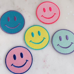 Puede incluir: Cinco parches bordados con caritas sonrientes de diferentes colores. Los parches son rosa, amarillo, verde azulado, verde claro y rosa oscuro. Cada parche tiene un dise&ntilde;o simple de carita sonriente con dos ojos y una boca curva.