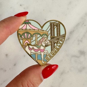 May include: Heart-shaped pin with a carousel and Brooklyn Bridge design. The pin features a carousel with a white horse, pink and green accents, and the word "BROOKLYN" in gold lettering. The background is light blue.