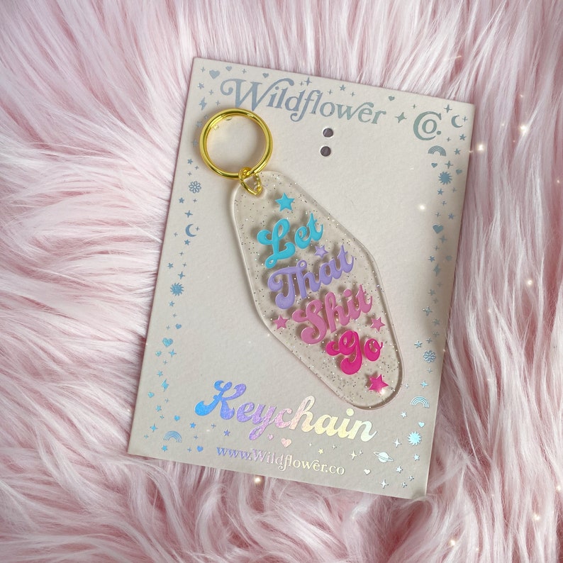 May include: Clear acrylic keychain with a gold ring, displaying the phrase "Let That Shit Go" in blue, purple, and pink. The keychain is on a card with the text "Wildflower & Co." and "Keychain".