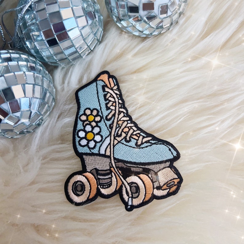 May include: Embroidered patch of a light blue roller skate with daisy accents. The patch features tan wheels and laces, with black outlines. The patch is set against a fluffy white background, with silver disco ball ornaments.
