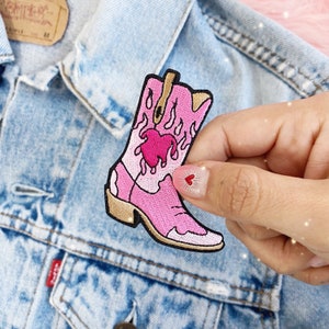May include: Pink cowboy boot embroidered patch with a dripping pink heart and flames. The patch is on a light blue denim jacket.