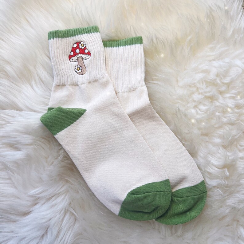K&ouml;nnte beinhalten: Ein Paar wei&szlig;e Kn&ouml;chelsocken mit gr&uuml;nem Besatz und einem roten und wei&szlig;en Pilz, der auf einer Socke gestickt ist.