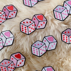 Könnte beinhalten: Ein Set mit 10 pinkfarbenen und weißen Filzwürfeln mit gestickten Herzen. Die Würfel sind in einem zufälligen Muster auf einer weißen, flauschigen Oberfläche angeordnet.