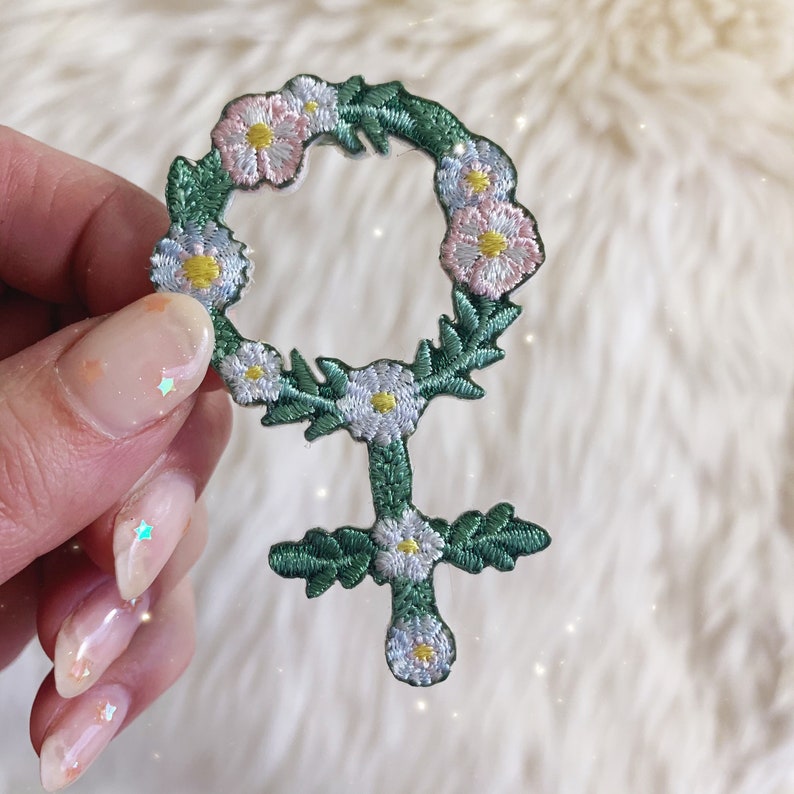 May include: A green and white embroidered patch in the shape of the female symbol. The symbol is surrounded by a wreath of white and pink flowers.