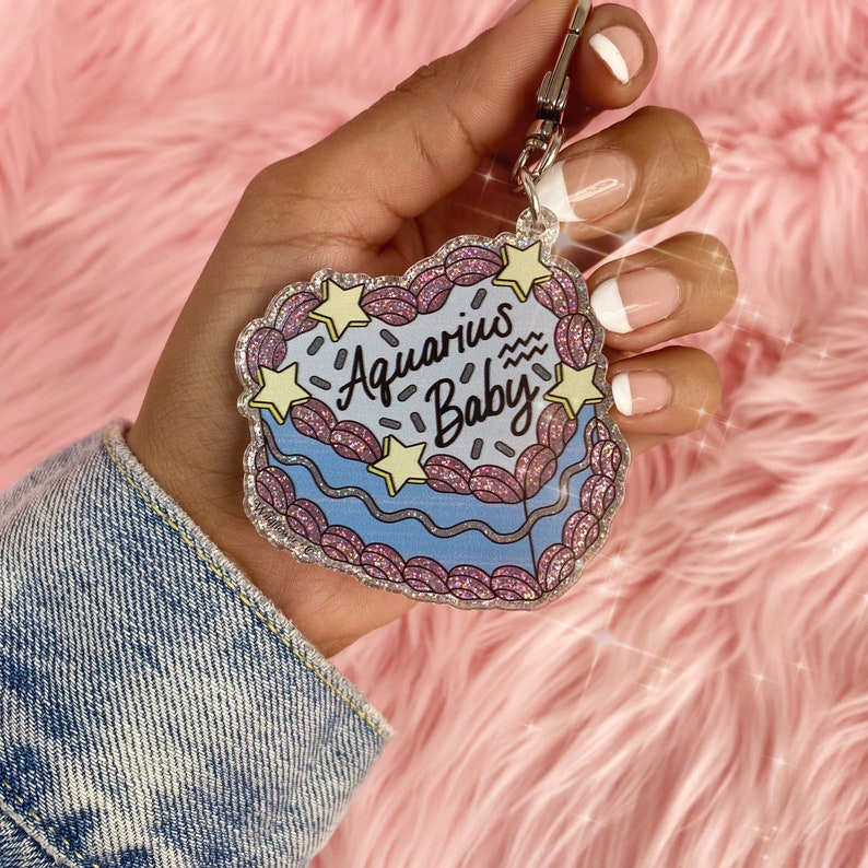 May include: A blue and pink glitter keychain with the text "Aquarius Baby" and a wavy line representing the astrological sign Aquarius.