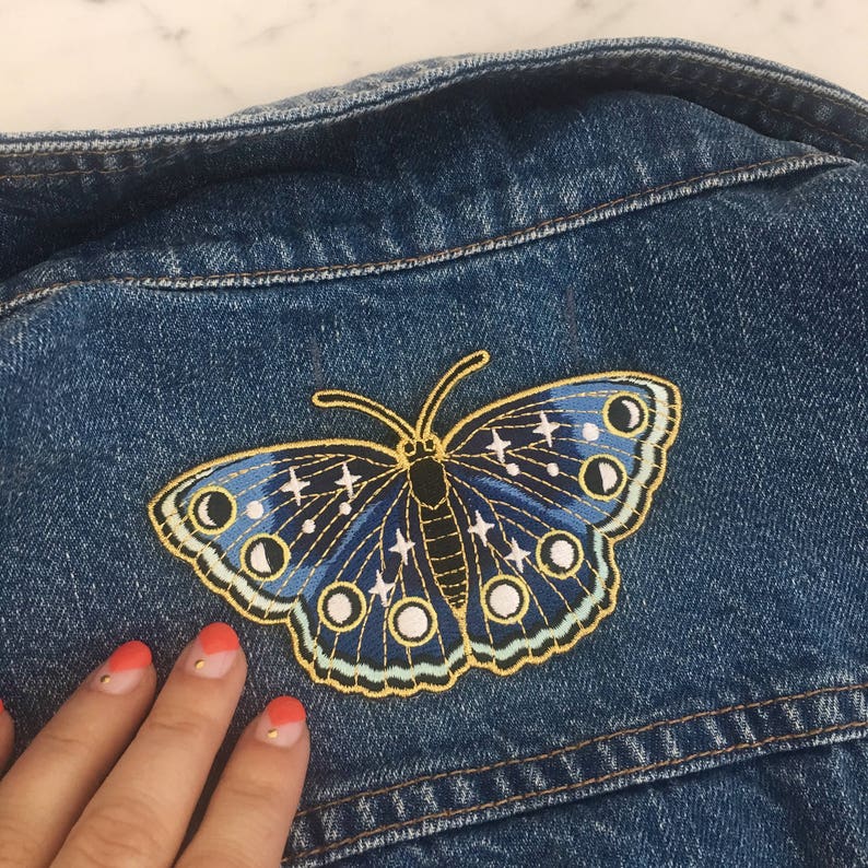 以下が含まれることがあります： 月と星のディテールが施された、青と金の刺繍の蝶のワッペン。ワッペンは青いデニムジャケットに縫い付けられています。