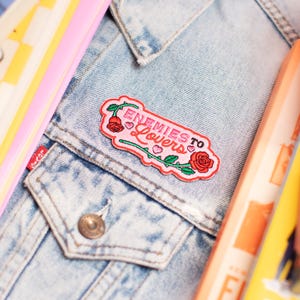 May include: Embroidered patch with the text "Enemies to Lovers" in pink and white, featuring a rose and vine design. The patch is attached to a light blue denim jacket. Other colourful items are in the background.