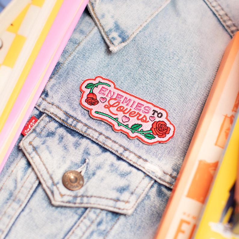 May include: Embroidered patch with the text "Enemies to Lovers" in pink and white, featuring a rose and vine design. The patch is attached to a light blue denim jacket. Other colourful items are in the background.