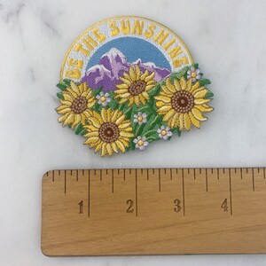 May include: A yellow and green embroidered patch with the text "Be the Sunshine" in a white circle. The patch features a mountain range and a cluster of yellow sunflowers.