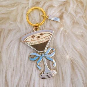 May include: A gold-coloured keyring with an espresso martini charm. The charm features a martini glass design with a light blue bow, brown coffee, and the text "Espresso Martini Club". A small tag with the text "Wildfire" is attached.