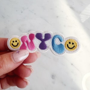 May include: A white felt patch with the letters NYC in colorful, embroidered, stylized font. The letters are surrounded by two yellow smiley faces.