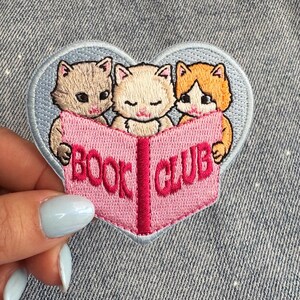 May include: Embroidered patch showing three kittens reading a pink book with the words "BOOK CLUB". The patch is heart-shaped with a light blue border. Ideal for book lovers and cat fans.