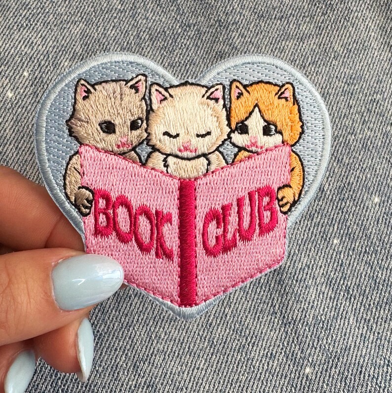 May include: Embroidered patch showing three kittens reading a pink book with the words "BOOK CLUB". The patch is heart-shaped with a light blue border. Ideal for book lovers and cat fans.