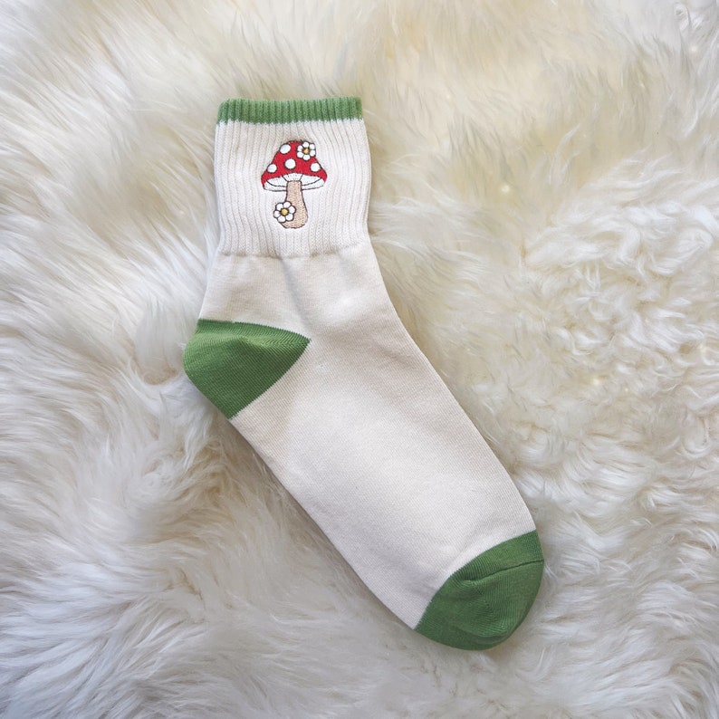 K&ouml;nnte beinhalten: Wei&szlig;e Kn&ouml;chelsocken mit gr&uuml;nem Saum und einem roten und wei&szlig;en Pilzmotiv. Der Pilz hat wei&szlig;e Punkte und eine wei&szlig;e Blume oben drauf.