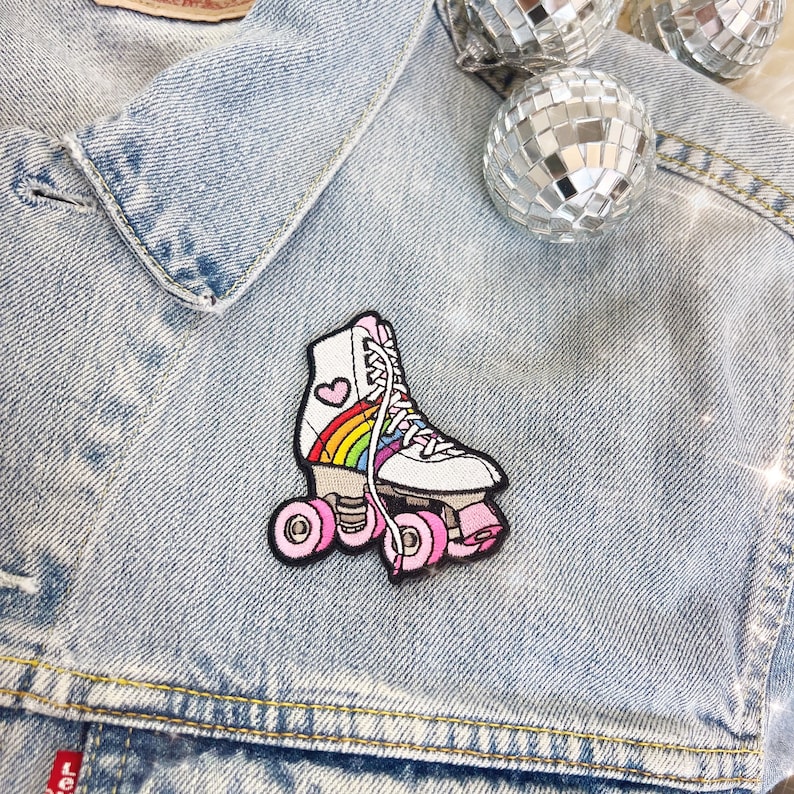 May include: A white roller skate patch featuring a rainbow and heart design. The patch has pink wheels and laces, and is on a light blue denim jacket. Disco balls are in the background.