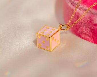 Puede incluir: Un collar de cadena dorada con un dije de dados rosa y dorado. El dado tiene corazones dorados en cada lado.
