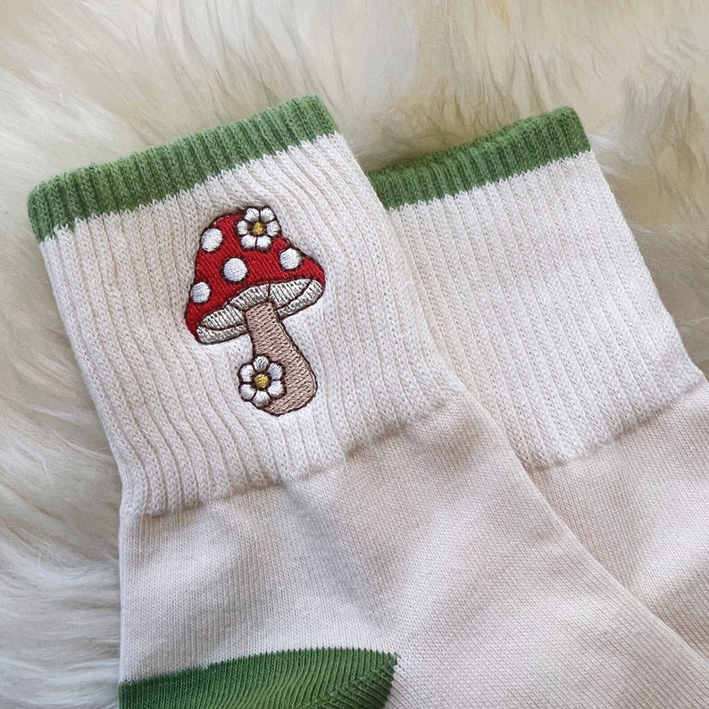 K&ouml;nnte beinhalten: Ein Paar wei&szlig;e Socken mit gr&uuml;nem Besatz und einem roten und wei&szlig;en Pilz, der auf einer Socke gestickt ist.
